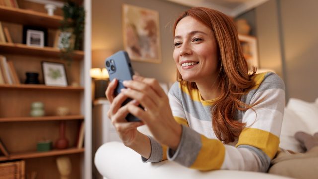 Rothaarige Frau liegt auf dem Sofa und schaut auf ihr iPhone