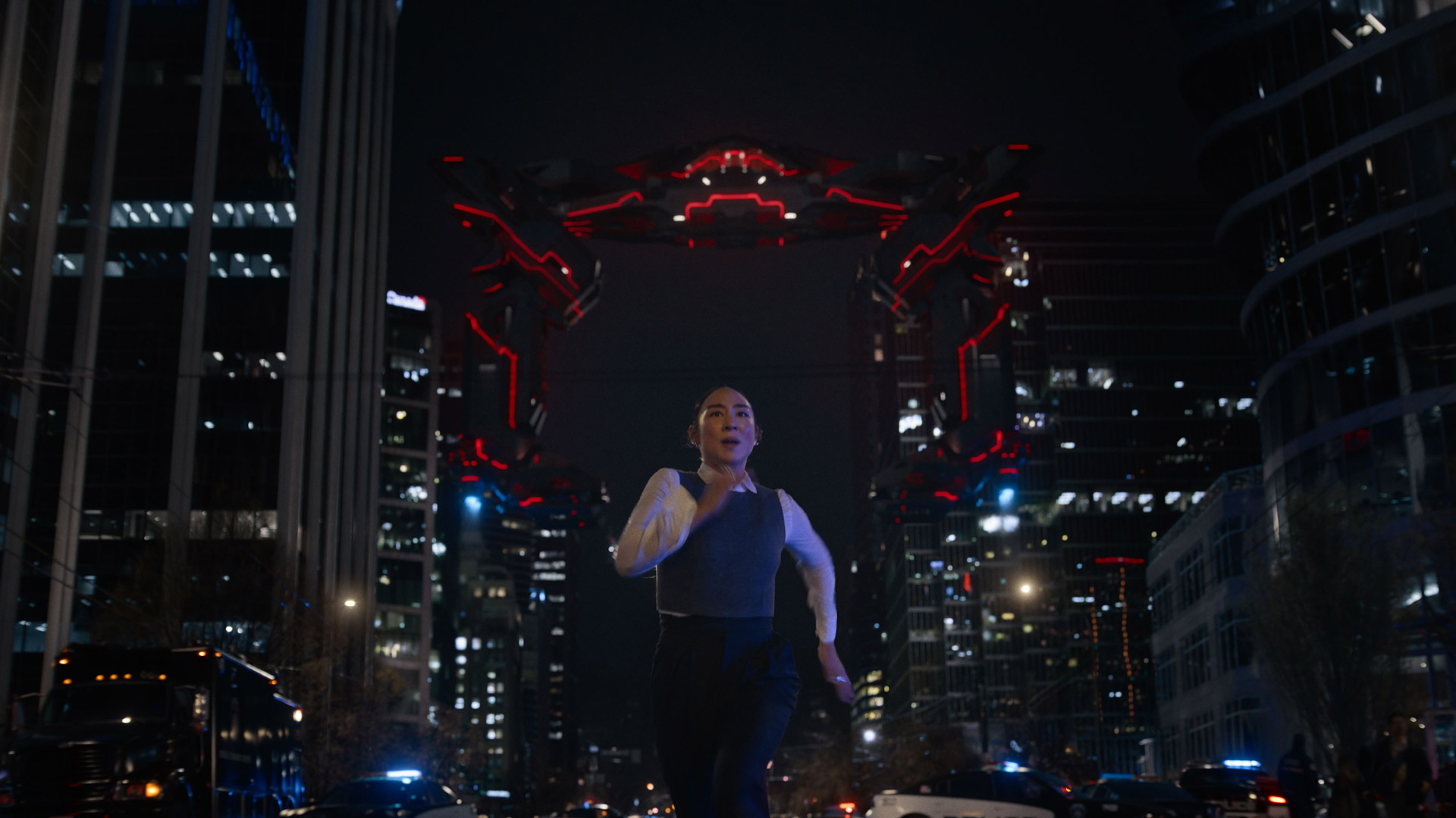 In dieser Szene aus dem Film Tron: Ares läuft Greta Lee als Eve Kim durch eine nächtliche Großstadt, sichtbar in Panik. Im Hintergrund erhebt sich ein riesiges, leuchtend rot illuminiertes Tor mit futuristischem Design zwischen zwei Hochhäusern.