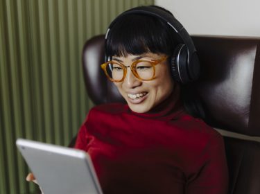 Frau mit Over-Ear-Kopfhörer sitzt auf einem Sessel und schaut auf ein Tablet.