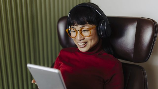 Frau mit Over-Ear-Kopfhörer sitzt auf einem Sessel und schaut auf ein Tablet.