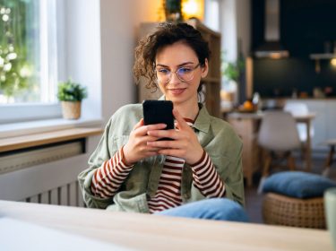 Junge Frau sitzt am Tisch und surft auf ihrem Handy.