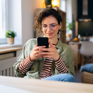 Junge Frau sitzt am Tisch und surft auf ihrem Handy.