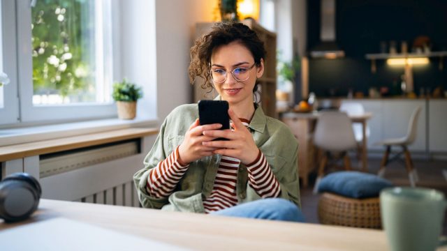 Junge Frau sitzt am Tisch und surft auf ihrem Handy.