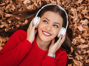 Eine Frau mit Over-Ear-Kopfhörern liegt auf einem Boden inmitten von herabgefallender Herbstblätter.