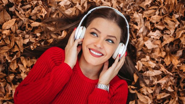 Eine Frau mit Over-Ear-Kopfhörern liegt auf einem Boden inmitten von herabgefallender Herbstblätter.