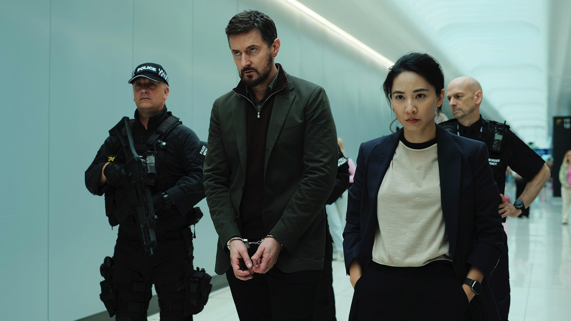In den GigaTV-Highlights im August ist eine Szene mit Dr. Matthew Nolan, gespielt von Richard Armitage, und einer Polizistin, dargestellt von Jing Lusi, zu sehen. Nolan wird mit Handschellen von bewaffneten Beamten abgeführt, während Lusi ernst und entschlossen voranschreitet.