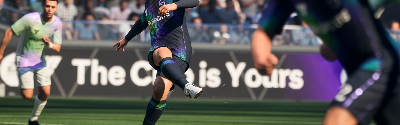 Auf diesem Screenshot aus dem Fußball-Videospiel „EA Sports FC 26“ ist eine Spielsituation auf dem Rasen zu sehen: Ein Spieler im dunkelblauen Trikot mit Regenbogenakzenten schießt konzentriert den Ball. Im Hintergrund sind Gegner und Zuschauer zu erkennen. Die Szene wirkt dynamisch und realitätsnah inszeniert.