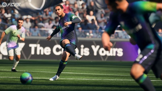Auf diesem Screenshot aus dem Fußball-Videospiel „EA Sports FC 26“ ist eine Spielsituation auf dem Rasen zu sehen: Ein Spieler im dunkelblauen Trikot mit Regenbogenakzenten schießt konzentriert den Ball. Im Hintergrund sind Gegner und Zuschauer zu erkennen. Die Szene wirkt dynamisch und realitätsnah inszeniert.