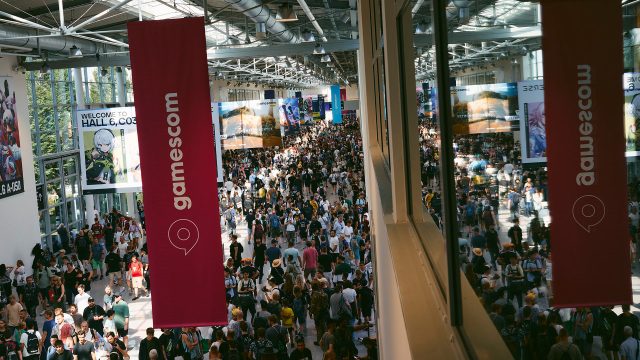 Auf dem Bild in dem Artikel zur gamescom 2025 ist eine Menschenmenge in einer hellen Messehalle zu sehen. Zwei große Banner mit der Aufschrift „gamescom“ hängen von der Decke.