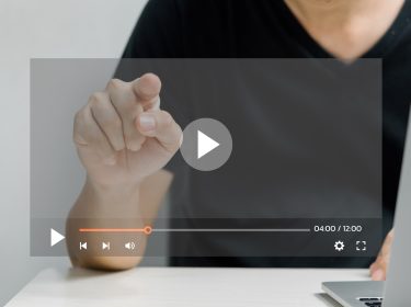 Ein Mann in schwarzem T-Shirt sitzt an einem Tisch und zeigt mit dem Finger auf einen transparent eingeblendeten Play-Button eines Videoplayers. Auf der rechten Seite ist ein aufgeklappter Laptop zu sehen. Das Bild symbolisiert die Nutzung eines digitalen Tools und steht stellvertretend für das Thema Sora Videos erstellen.