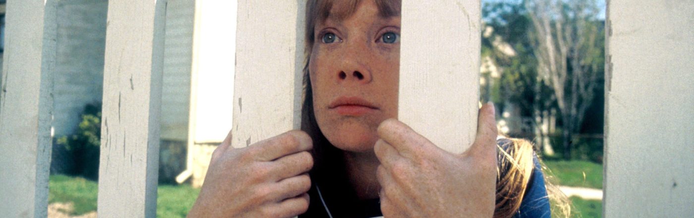 Carrie schaut durch einen Lattenzaun. Sie kniet und hält sich daran fest.