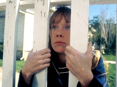 Carrie schaut durch einen Lattenzaun. Sie kniet und hält sich daran fest.