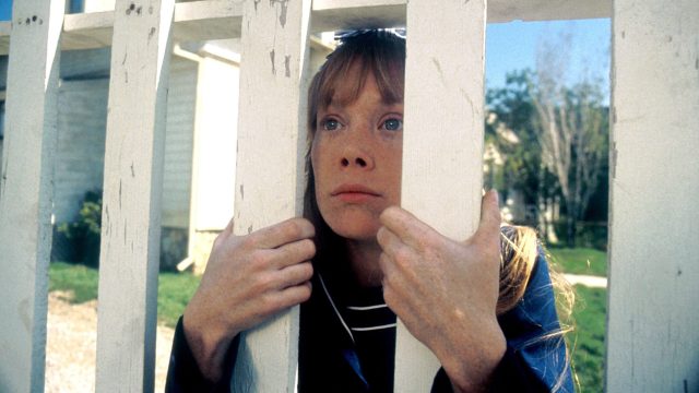 Carrie schaut durch einen Lattenzaun. Sie kniet und hält sich daran fest.