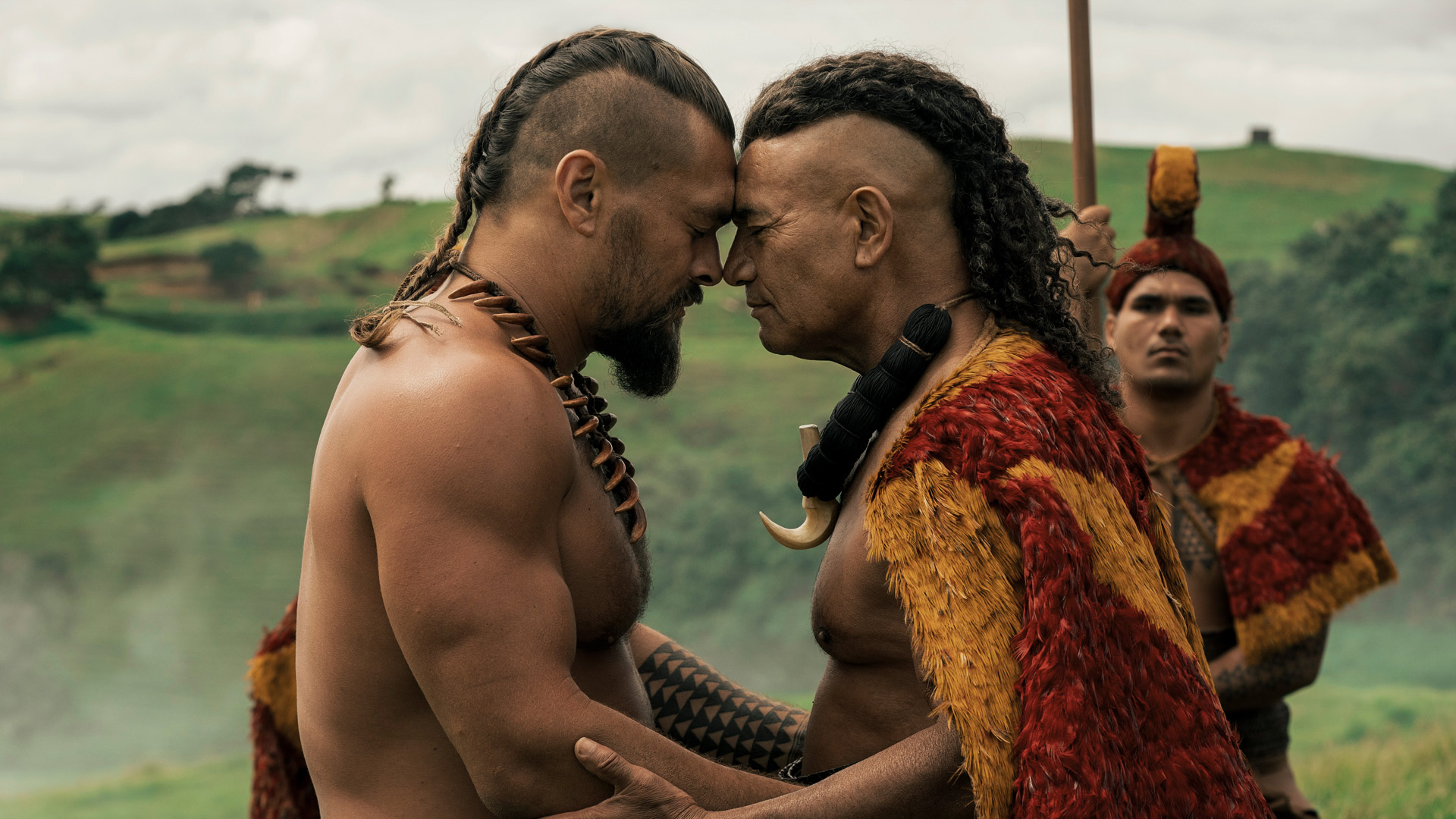 Auf dem Bild aus Chief of War sind Jason Momoa (links) und Temuera Morrison (rechts) in einer emotionalen Szene zu sehen. Beide Männer stehen Stirn an Stirn, eine Geste des Respekts und der Verbundenheit. Sie tragen traditionelle Kleidung und stehen in einer grünen Hügellandschaft. Die Szene wirkt würdevoll und bedeutungsvoll. 
