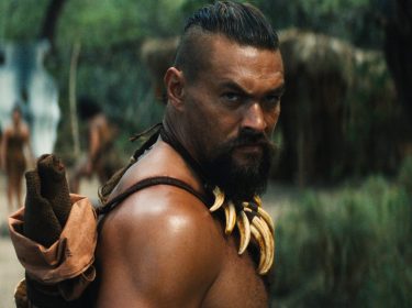 Auf dem Bild aus Chief of War ist Jason Momoa in einer intensiven Szene zu sehen. Er trägt eine Halskette aus Tierzähnen und blickt ernsthaft über die Schulter. Im Hintergrund stehen weitere Stammesmitglieder in traditioneller Kleidung in einer bewaldeten Umgebung. Die Szene wirkt angespannt und eindrucksvoll.