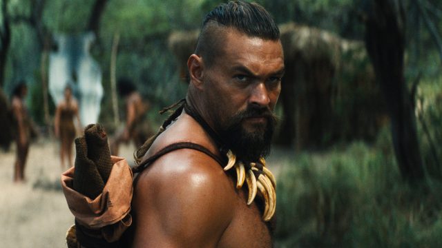 Auf dem Bild aus Chief of War ist Jason Momoa in einer intensiven Szene zu sehen. Er trägt eine Halskette aus Tierzähnen und blickt ernsthaft über die Schulter. Im Hintergrund stehen weitere Stammesmitglieder in traditioneller Kleidung in einer bewaldeten Umgebung. Die Szene wirkt angespannt und eindrucksvoll.