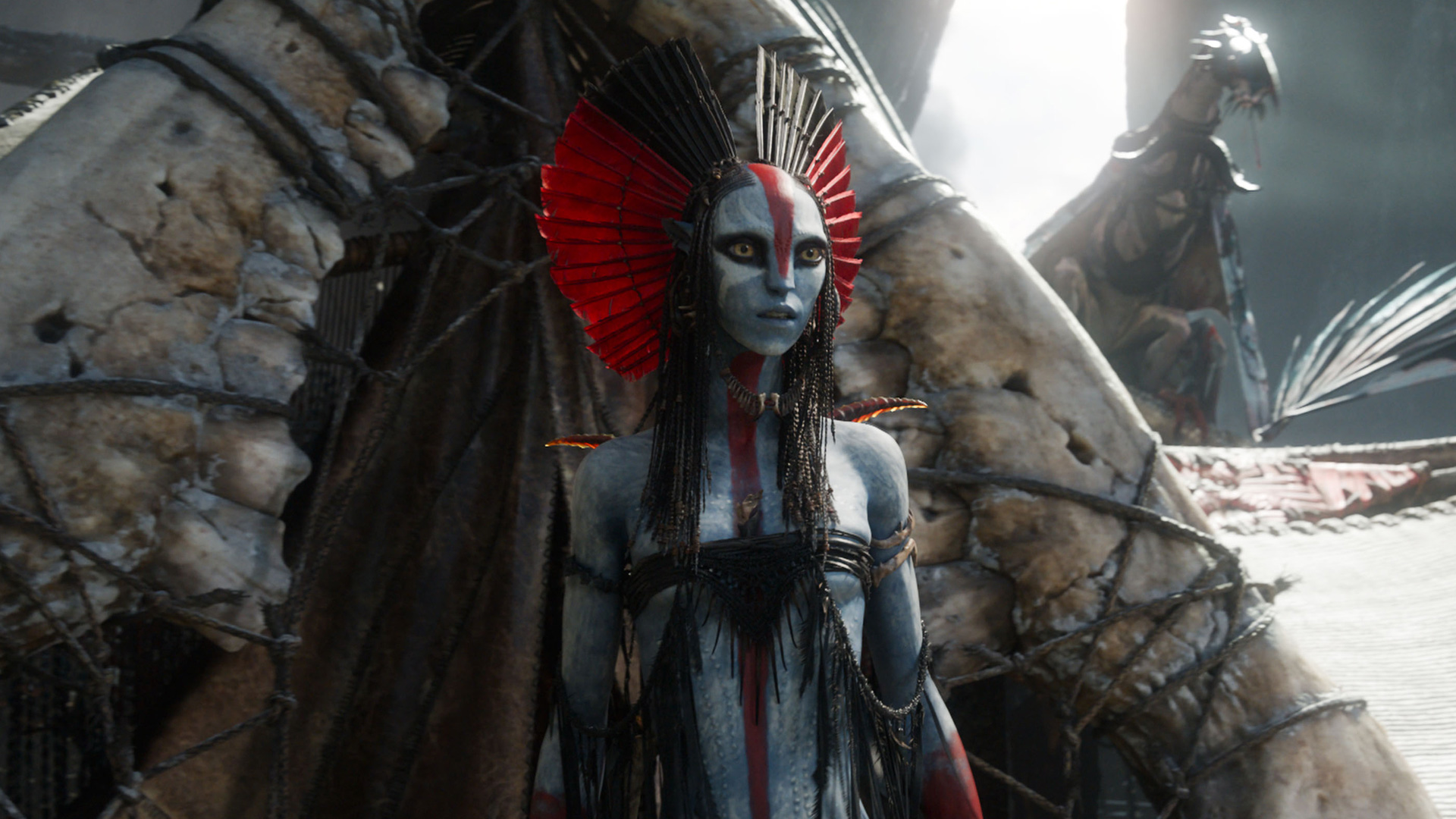 Auf dem Bild aus Avatar 3 ist eine weibliche Na’vi mit blauer Haut und auffälliger rot-schwarzer Kopfbedeckung zu sehen. Ihr Gesicht ist mit einem roten Streifen bemalt.