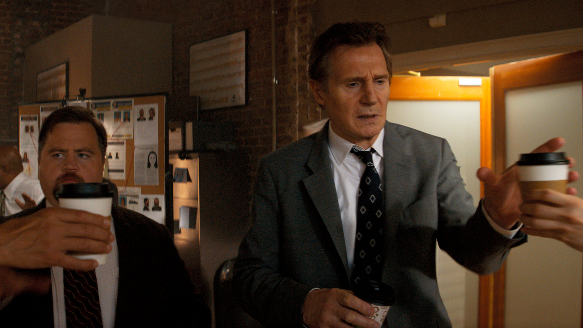 In einer Szene aus Die nackte Kanone sind Paul Walter Hauser als Ed Hocken Jr. und Liam Neeson als Frank Drebin Jr. in einem hektischen Büro zu sehen. Beide tragen Anzug und halten Kaffeebecher, während sie sich angespannt durch den Raum bewegen. Die Szene vermittelt eine Mischung aus Chaos und Komik – typisch für Die nackte Kanone.