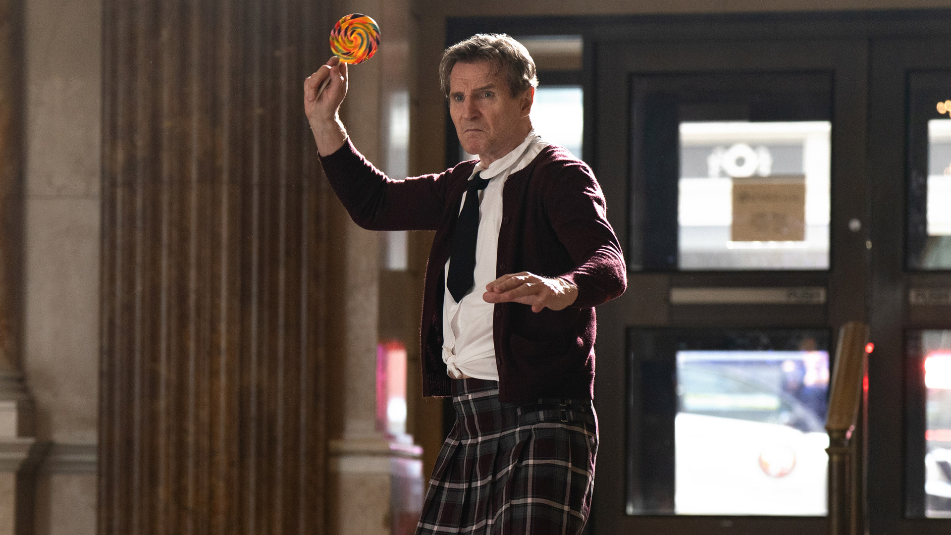 Liam Neeson ist in der Rolle des Frank Drebin Jr. in einer absurden Actionszene aus dem neuen Die nackte Kanone Film zu sehen. In angespannter Kampfpose hält er einen riesigen Lolli wie eine Waffe. Er trägt ein zerzaustes Hemd, Krawatte, Strickjacke und einen karierten Kilt – eine typische Mischung aus Slapstick und Ernst, wie sie für Die nackte Kanone charakteristisch ist.