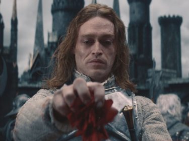 In einer Schlüsselszene aus Dracula - Die Auferstehung steht Dracula, gespielt von Caleb Landry Jones, in silberner Rüstung vor einer düsteren Burgkulisse. Mit ernstem Blick hält er eine rote Blume in der Hand, als Symbol für Verlust oder Erinnerung. Der Himmel ist grau, die Stimmung bedrohlich und melancholisch zugleich.