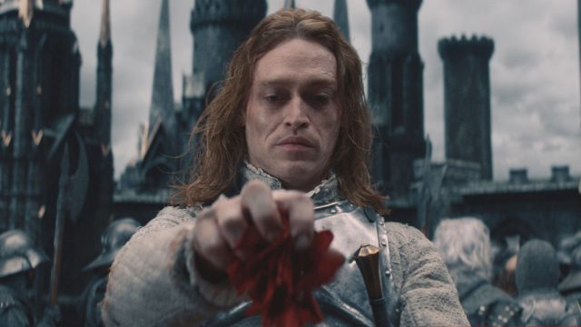 In einer Schlüsselszene aus Dracula - Die Auferstehung steht Dracula, gespielt von Caleb Landry Jones, in silberner Rüstung vor einer düsteren Burgkulisse. Mit ernstem Blick hält er eine rote Blume in der Hand, als Symbol für Verlust oder Erinnerung. Der Himmel ist grau, die Stimmung bedrohlich und melancholisch zugleich.