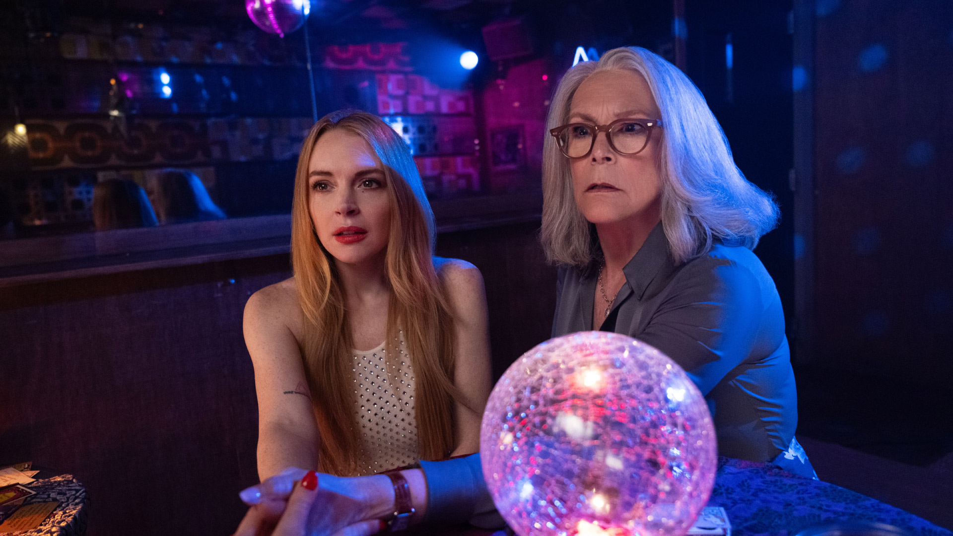 In dieser Szene aus Freakier Friday sitzen Jamie Lee Curtis als Tess Coleman und Lindsay Lohan als Anna Coleman nebeneinander in einer dunklen, stimmungsvoll beleuchteten Bar. Beide blicken gespannt und etwas besorgt nach vorn, vor ihnen liegt eine glitzernde Kristallkugel. Die Atmosphäre wirkt geheimnisvoll und leicht surreal – passend für eine Freakier Friday Kritik