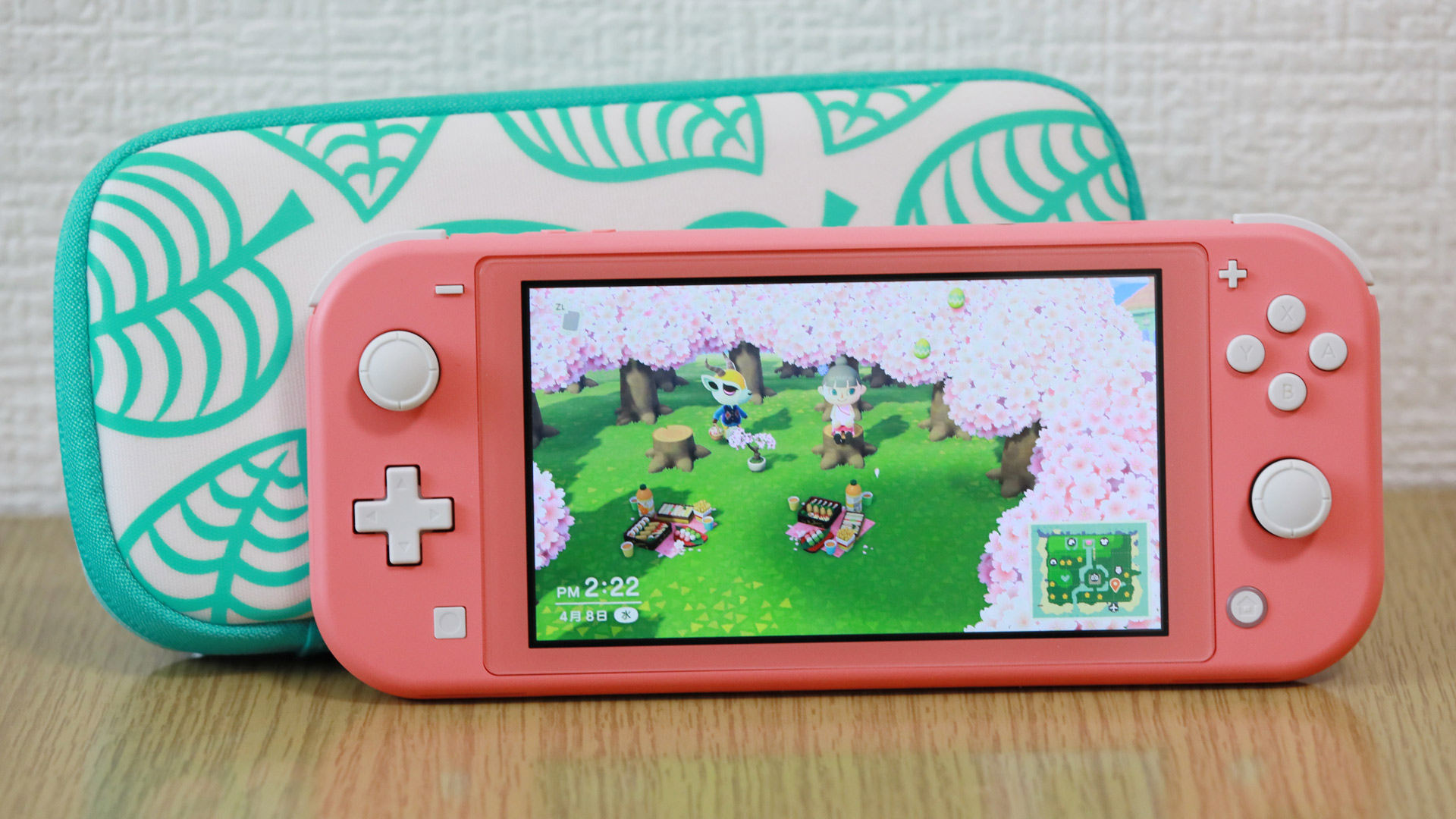 Auf dem Bild ist eine pinke Nintendo Switch Lite zu sehen, auf der das Spiel Animal Crossing: New Horizons läuft. Zwei Spielfiguren sitzen beim Picknick unter Kirschblütenbäumen. Im Hintergrund liegt ein passendes Etui mit Blattmuster. Das Bild dient symbolisch zur Illustration eines möglichen neuen Animal Crossing Switch 2 Titels.