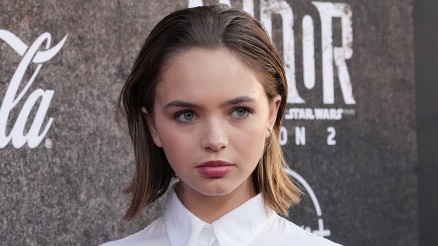 Auf dem Bild in dem Artikel zum Buffy Reboot ist Schauspielerin Ryan Kiera Armstrong zu sehen. Sie steht vor einer dunklen Fotowand, trägt ein weißes Hemd und blickt ernst in die Kamera.