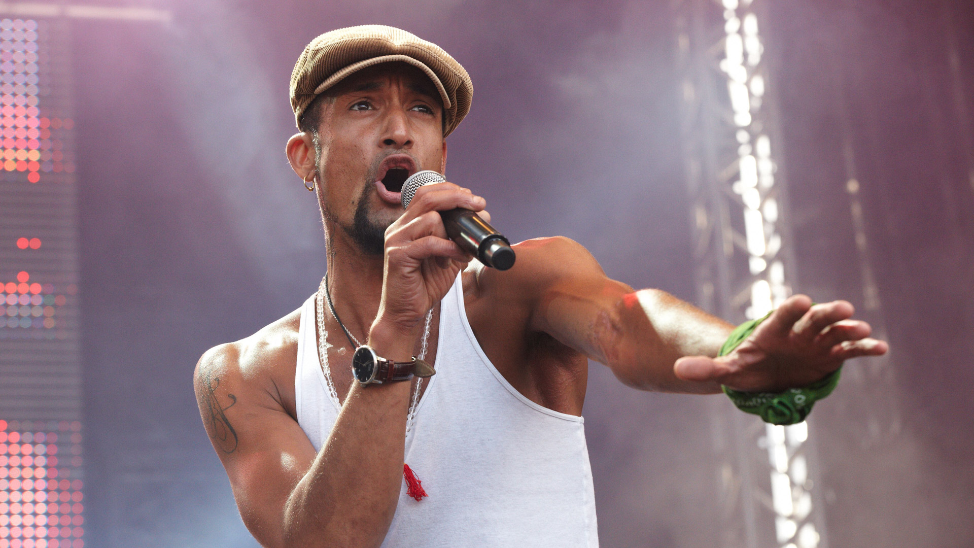 Auf dem Bild ist DSDS-Gewinner Mark Medlock bei einem Live-Auftritt zu sehen. Er trägt ein weißes Tanktop, eine braune Schirmmütze und hält ein Mikrofon in der Hand, während er leidenschaftlich singt. Mit ausdrucksstarker Mimik und ausgestrecktem Arm verkörpert er die Energie und das Charisma erfolgreicher DSDS-Gewinner:innen auf der Bühne.