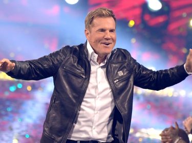 Auf dem Bild ist Dieter Bohlen mit ausgebreiteten Armen und einem breiten Lächeln zu sehen. Er trägt eine schwarze Lederjacke und steht auf einer Bühne mit bunten Lichtern und Konfetti im Hintergrund – die Stimmung ist ausgelassen und feierlich. Als langjähriges Gesicht der Show steht Bohlen symbolisch für die DSDS-Gewinner:innen.