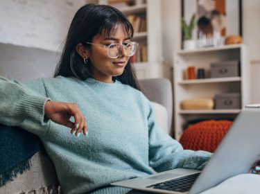 Auf dem Bild sitzt eine junge Frau mit langem dunklem Haar und Brille entspannt auf einem Sofa und arbeitet an einem Laptop. Sie trägt einen hellblauen Pullover und wirkt konzentriert. Das Wohnzimmer im Hintergrund ist modern eingerichtet. Die Szene symbolisiert den alltäglichen Umgang mit ChatGPT-Modelle im privaten Umfeld.