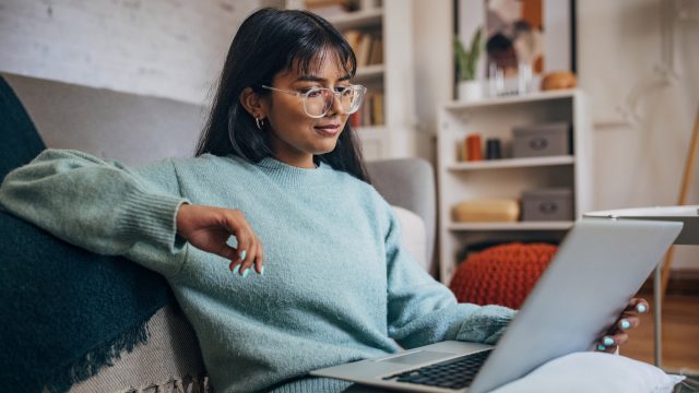 Auf dem Bild sitzt eine junge Frau mit langem dunklem Haar und Brille entspannt auf einem Sofa und arbeitet an einem Laptop. Sie trägt einen hellblauen Pullover und wirkt konzentriert. Das Wohnzimmer im Hintergrund ist modern eingerichtet. Die Szene symbolisiert den alltäglichen Umgang mit ChatGPT-Modelle im privaten Umfeld.