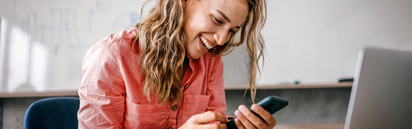 iPhone WLAN Hotspot einrichten: Frau sitzt lachend mit ihrem Smartphone in der Hand vor ihrem Laptop.