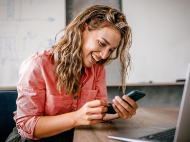iPhone WLAN Hotspot einrichten: Frau sitzt lachend mit ihrem Smartphone in der Hand vor ihrem Laptop.