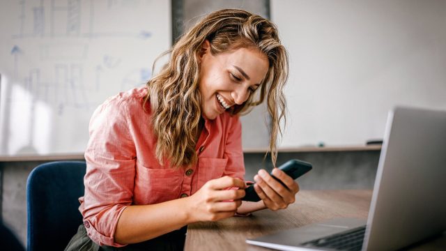 iPhone WLAN Hotspot einrichten: Frau sitzt lachend mit ihrem Smartphone in der Hand vor ihrem Laptop.