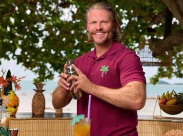 Das Bild zu Bachelor in Paradise 2025 zeigt Paul Janke, der an einer Außenbar steht. Er trägt ein bordeauxrotes Polohemd mit einem grünen Palmen-Logo und lächelt in die Kamera.