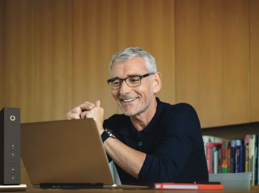 Vodafone Router einrichten: Mann mittleren Alters sitzt lächelnd vor seinem Laptop und Router.