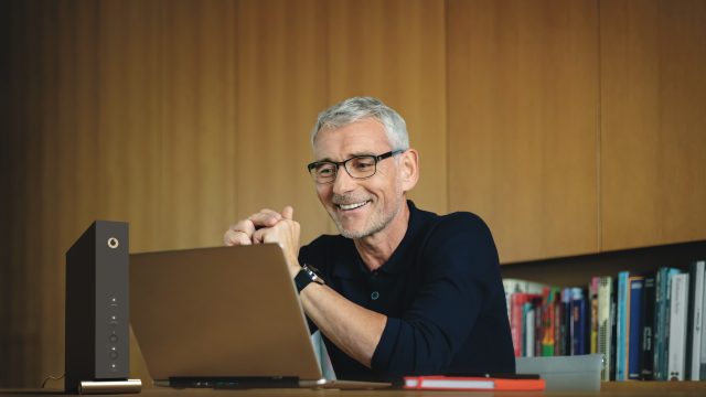 Vodafone Router einrichten: Mann mittleren Alters sitzt lächelnd vor seinem Laptop und Router.