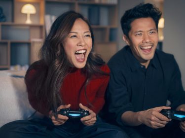 PS5 WLAN Probleme: Ein Mann und eine Frau sitzen aufgeregt auf einem Sofa und halten PlayStation Controller in der Hand.