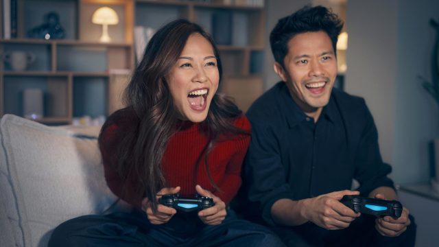 PS5 WLAN Probleme: Ein Mann und eine Frau sitzen aufgeregt auf einem Sofa und halten PlayStation Controller in der Hand.