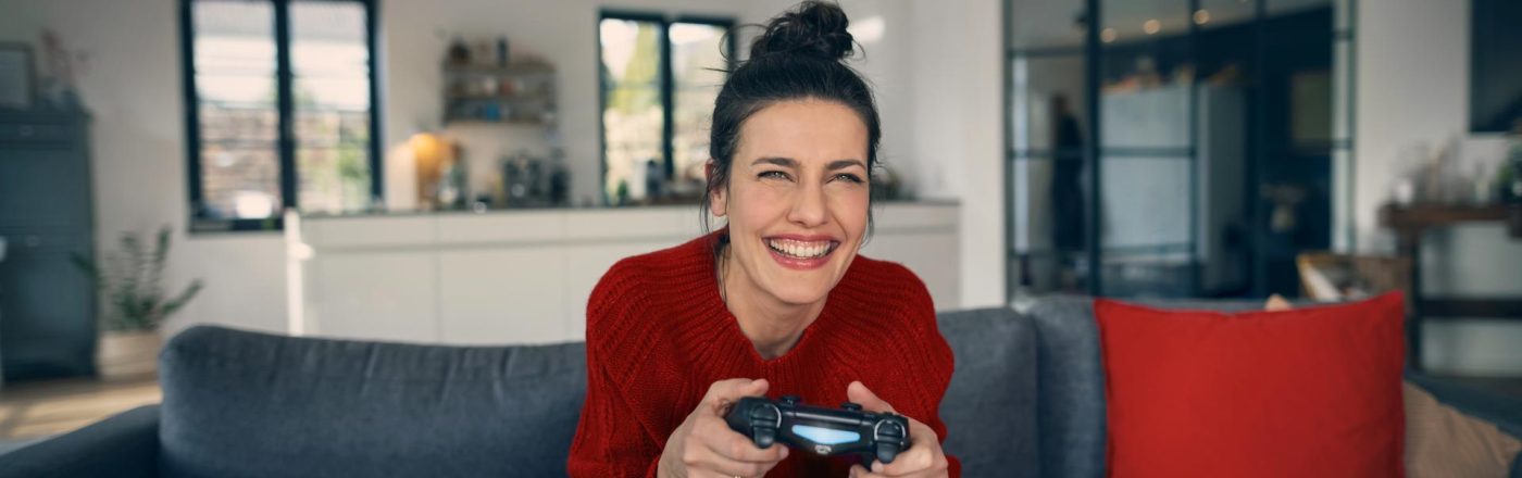 PS5 Upload Probleme: Frau sitzt freudig auf einem Sofa und hat einen PlayStation-Controller in der Hand.