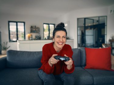 PS5 Upload Probleme: Frau sitzt freudig auf einem Sofa und hat einen PlayStation-Controller in der Hand.
