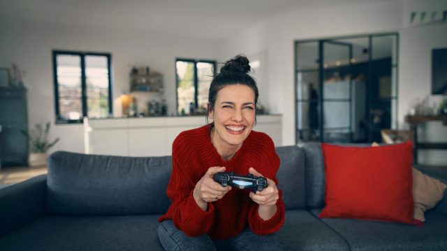 PS5 Upload Probleme: Frau sitzt freudig auf einem Sofa und hat einen PlayStation-Controller in der Hand.