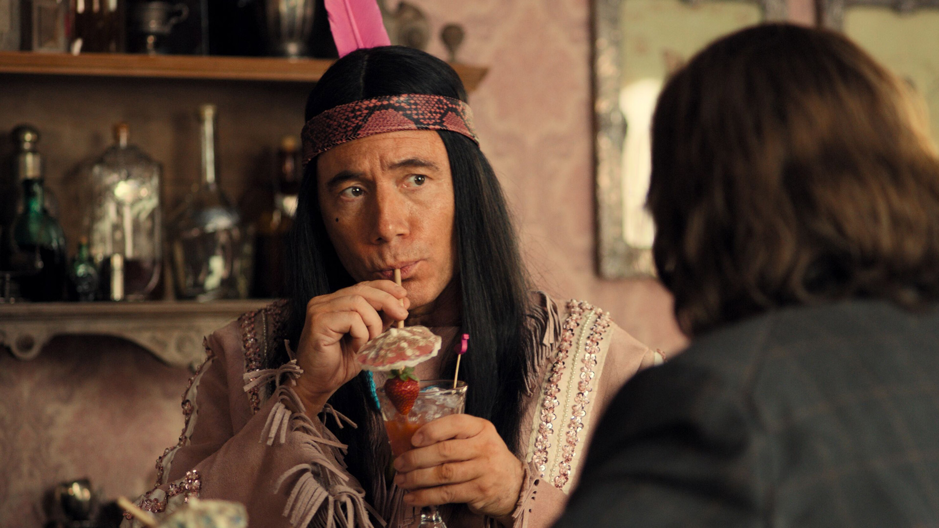 Auf dem Bild zur Das Kanu des Manitu Kritik ist Michael Bully Herbig in seiner Rolle als Winnetouch zu sehen. Er trägt ein glitzernd verziertes Outfit mit pinker Feder und trinkt genüsslich einen Cocktail mit Schirmchen und Erdbeere. Sein Blick ist aufmerksam, während ihm eine Person mit langen Haaren gegenübersitzt. Die Szene wirkt humorvoll und leicht ironisch.