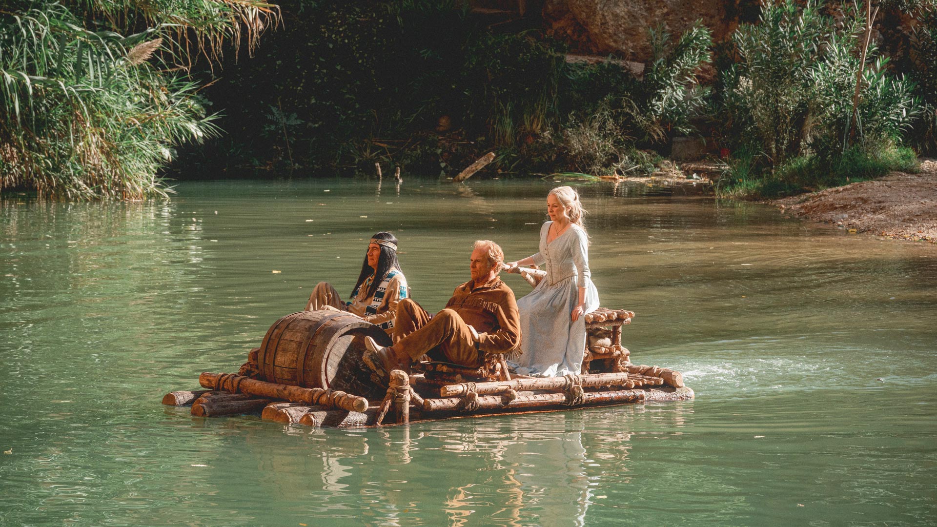 Für die Das Kanu des Manitu Kritik zeigt dieses Bild Abahachi (Michael Bully Herbig), Ranger (Christian Tramitz) und Mary (Jasmin Schwiers) auf einem selbstgebauten Floß aus Holzstämmen. Die drei Figuren befinden sich auf einem ruhigen Fluss, umgeben von dichtem Grün. Abahachi und Ranger sitzen vorn, während Mary am hinteren Ende des Floßes steht. Die Szene wirkt idyllisch und abenteuerlich.