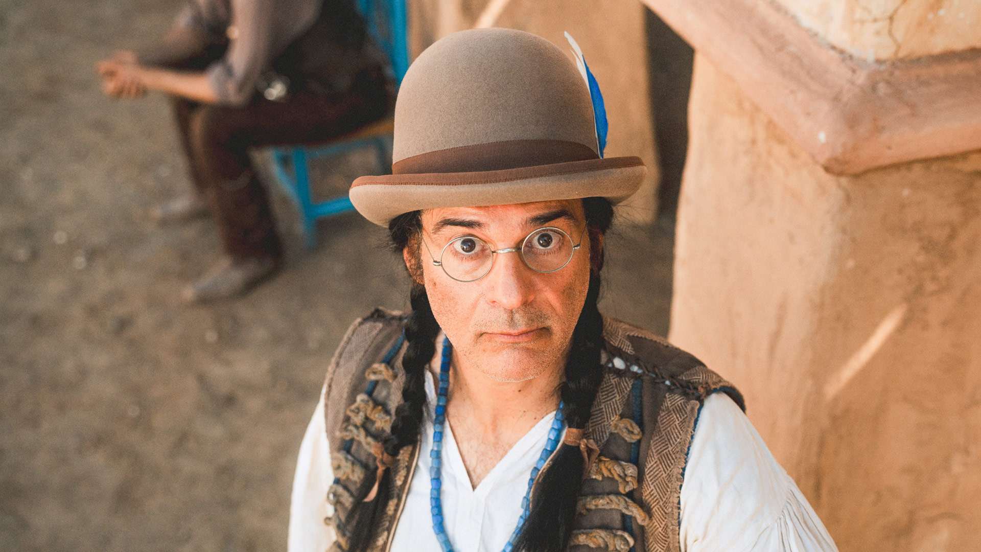 Auf dem Bild zur Das Kanu des Manitu Kritik ist Rick Kavanian in seiner Rolle als Dimitri zu sehen. Er blickt mit ernster Miene und großen Augen direkt in die Kamera. Dimitri trägt eine runde Brille, einen Filzhut mit Feder sowie zwei geflochtene Zöpfe. Im Hintergrund sitzt unscharf eine weitere Person auf einem blauen Stuhl.