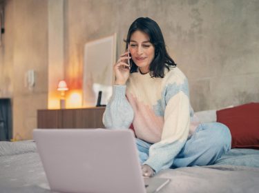 Vodafone Kabel-Internet- & Telefon-Anschluss freischalten: Frau sitzt telefonierend vor ihrem Laptop auf dem Bett.