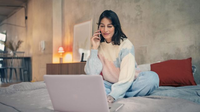 Vodafone Kabel-Internet- & Telefon-Anschluss freischalten: Frau sitzt telefonierend vor ihrem Laptop auf dem Bett.