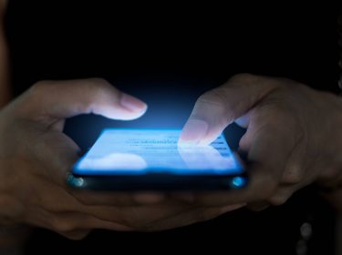 Nahaufnahme von zwei Händen, die in dunkler Umgebung auf einem Smartphone tippen.