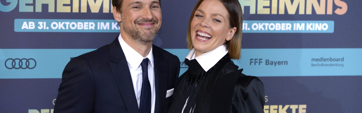 Auf diesem Bild zur symbolischen Ankündigung von Der perfekte Urlaub sind Florian David Fitz und Jessica Schwarz bei der Premiere von Das perfekte Geheimnis zu sehen. Beide Schauspieler lächeln herzlich in die Kamera. Fitz trägt einen dunklen Anzug mit Krawatte, während Schwarz mit weißem Hemd und schwarzem Blazer auffällt. Die Stimmung wirkt gelöst und fröhlich.